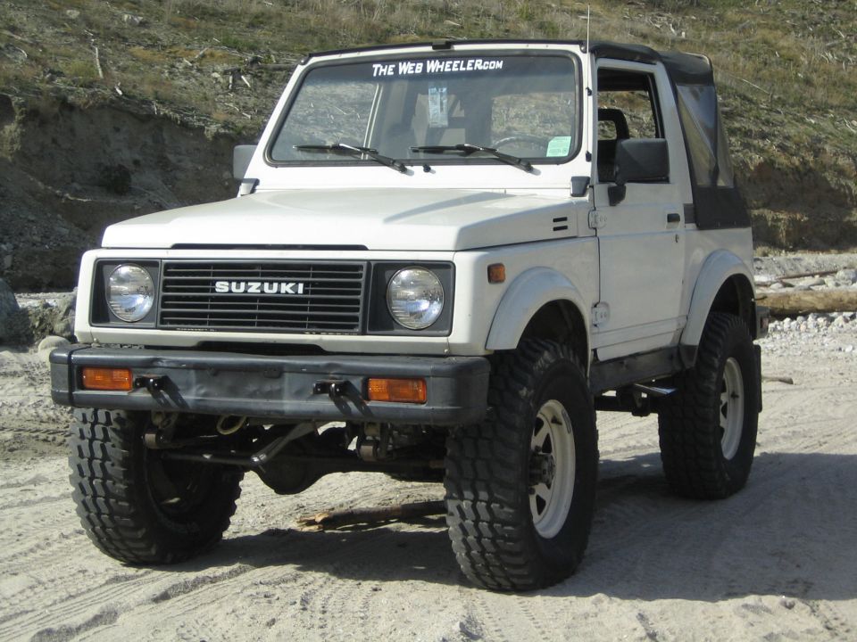 White 1987 Suzuki Samurai with black soft top, off-road tires, and a "The Web Wheeler.com" sticker on the windshield