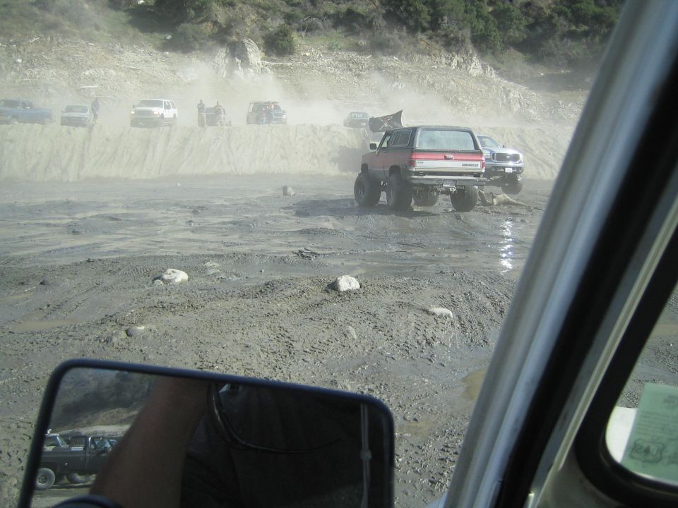 Red SUV with lifted suspension driving off-road in a dusty, muddy area with other vehicles
