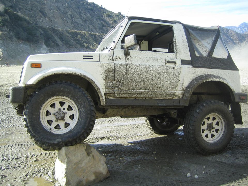 White Suzuki Samurai with mud splatters on its side, driving over a rock on a dirt road