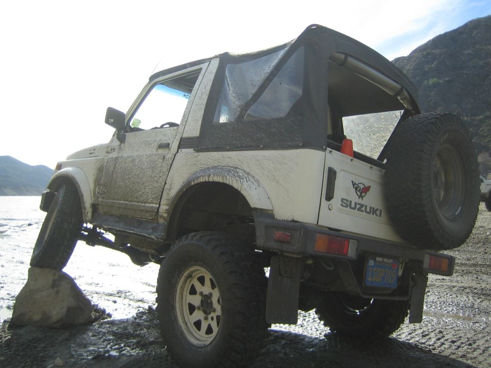 White 1987 Suzuki Samurai with soft top, rear-mounted spare tire, and off-road tires on a rocky trail