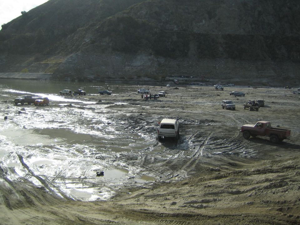 Red pickup truck and white van in muddy terrain with other vehicles in background