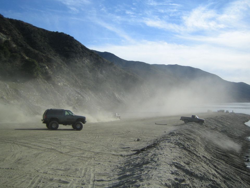 Several off-road vehicles driving on a dusty dirt road next to a lake with mountains in the background