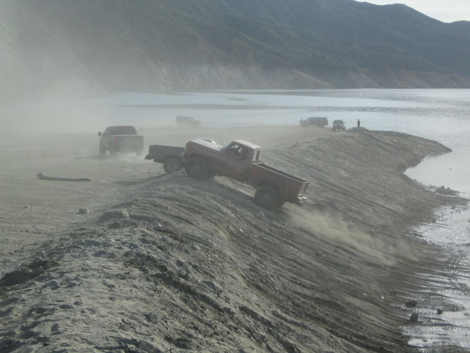 Red pickup truck driving on a dusty, dry lakebed near the water