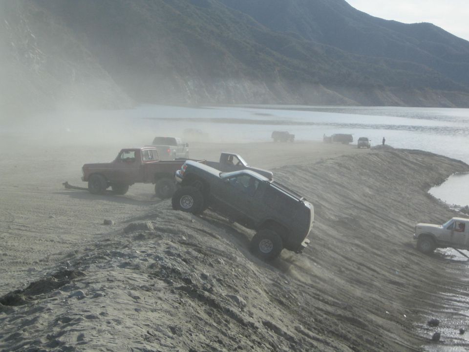 Dark pickup truck climbing dusty hill near water, other trucks visible