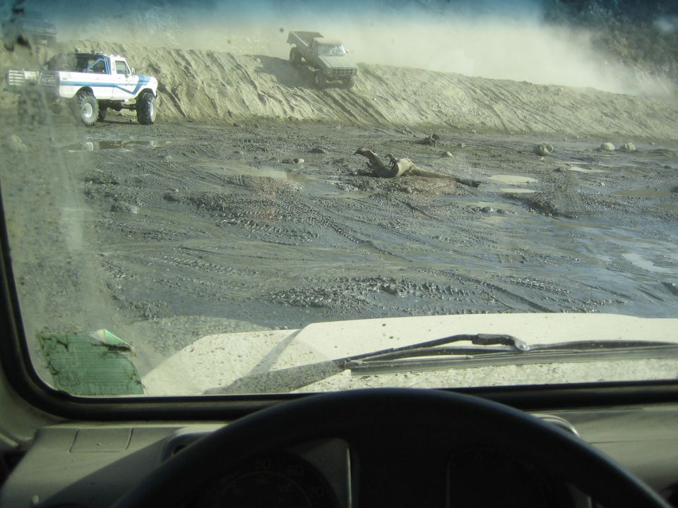 Two off-road trucks driving on a dusty dirt hill with mud and puddles below