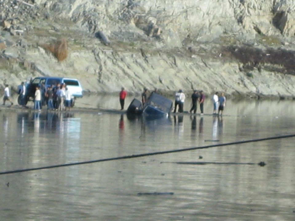 Two dark vehicles overturned in shallow water near a rocky shore, with a blue SUV and a group of people on the bank