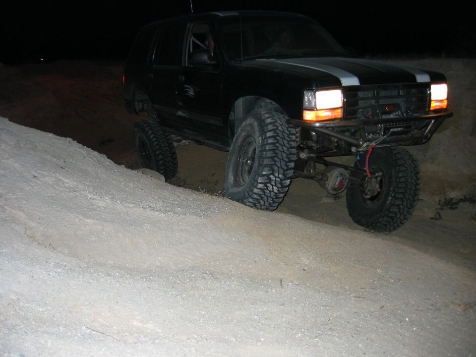 Modified black SUV with off-road tires climbing a dirt hill at night. Has racing stripes on hood and custom bumper