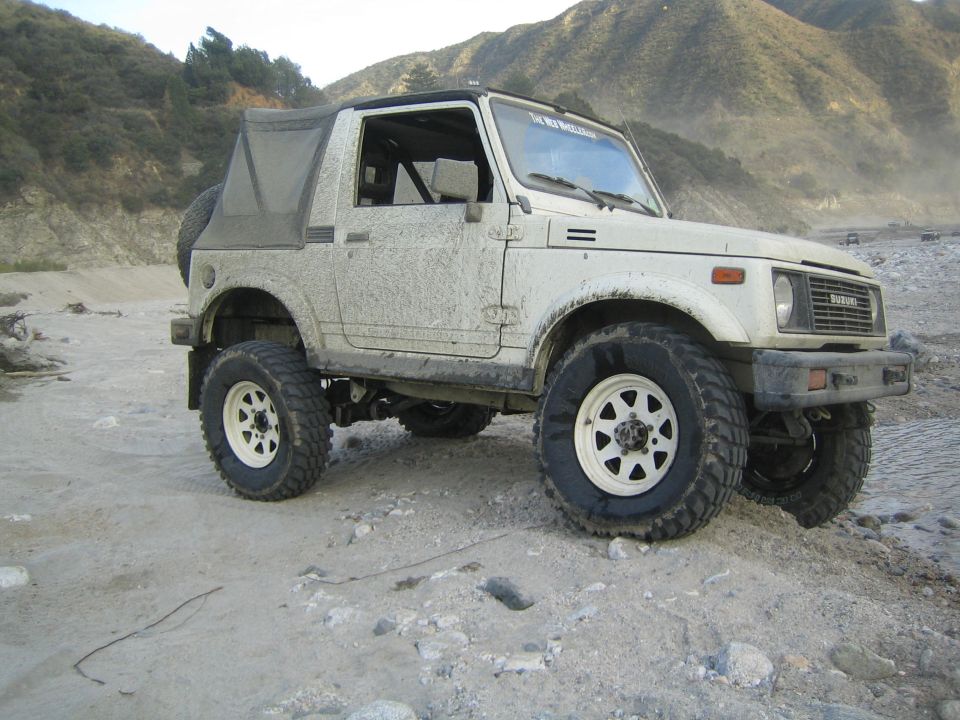 White Suzuki Samurai with soft top, covered in mud, on a dirt road with mountains in the background