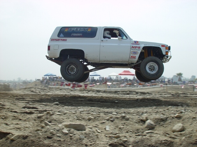 White off-road racing truck mid-air over dirt track. It has "FOX" and "DYNA TRAC" decals. The year appears to be around 1973