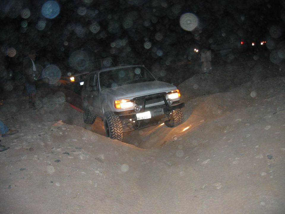 Lifted silver SUV flexing through a dirt rut at night with headlights on and dust in air