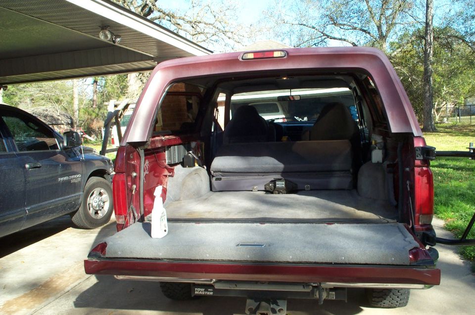 Red Ford Bronco rear cargo area open, showing gray carpet and folded seats. A blue pickup truck is parked to the left