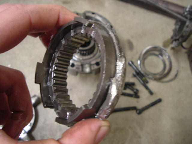 Hand holding a broken internally splined ring gear with a torn section, disassembled parts on floor