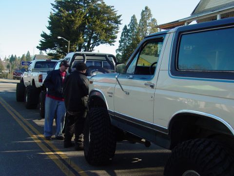 1986 Ford Bronco - A line of modified trucks and a Ford Bronco are parked on a street on a sunny day.