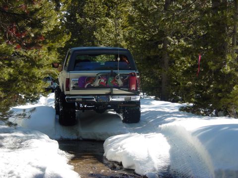 1986 Ford Bronco - A lifted Ford Bronco navigates a snowy path with snowboards attached to the rear.