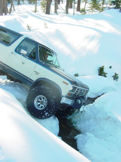 1986 Ford Bronco - A Ford Bronco drives through a snowy landscape, its tires deep in snow and water.