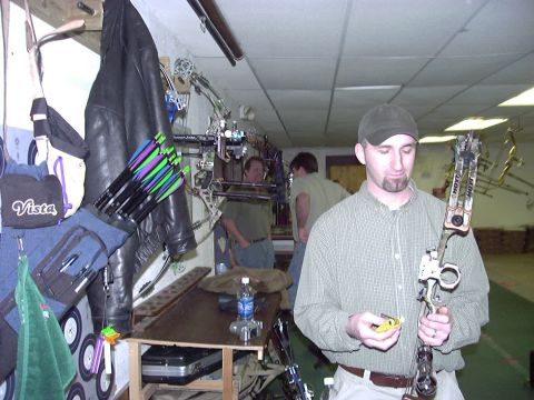 1992 Ford Bronco - A man inspects a compound bow in what appears to be an indoor archery range, with other people and archery equipment visible.
