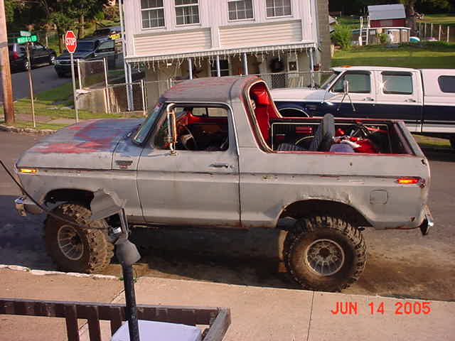 Gray primered Ford Bronco, top removed, red interior, lifted on mud tires, side view in driveway