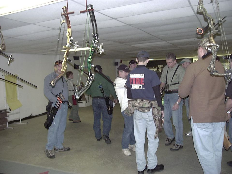 People gathered indoors with compound bows hanging from the ceiling