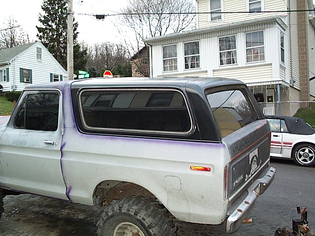 Gray primer Ford Bronco with purple stripes and black top, lifted on big tires, rear three-quarter view on a street