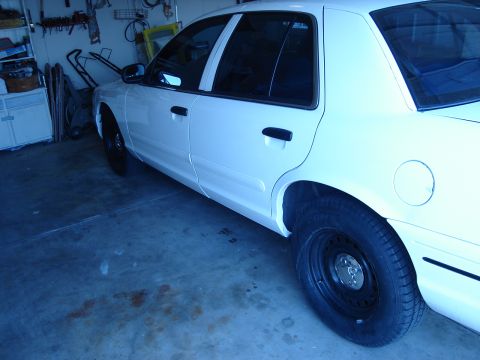 2000 Ford Crown Victoria - A white sedan with black steel wheels is parked inside a garage, showing its side profile.
