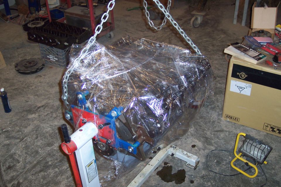 Engine wrapped in plastic, suspended by chains and resting on an engine stand