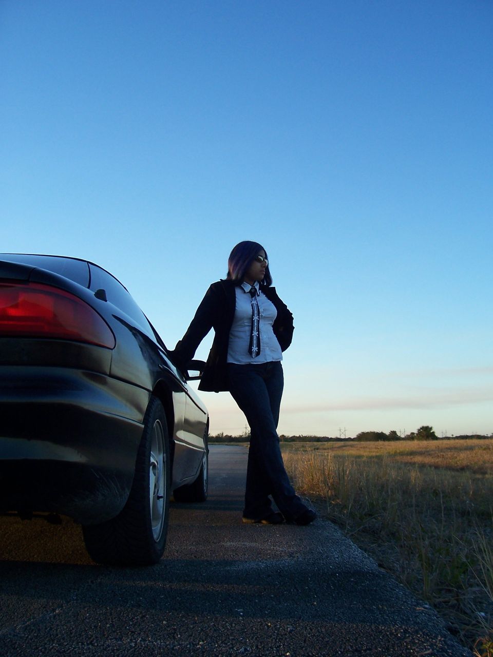 Woman in a suit jacket and tie leans against the rear quarter panel of a black car. The sky is clear blue