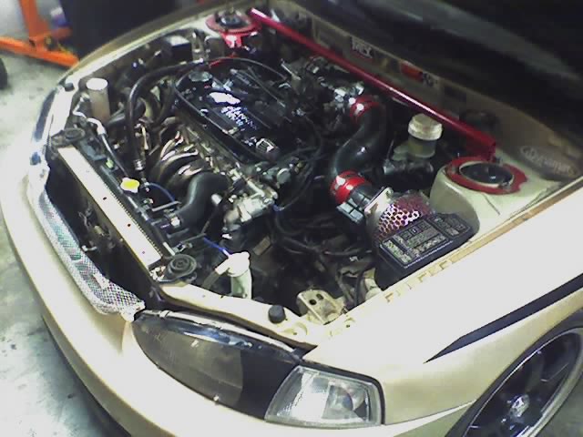 Gold compact car engine bay with exhaust header, red strut bar, and mesh cone intake filter