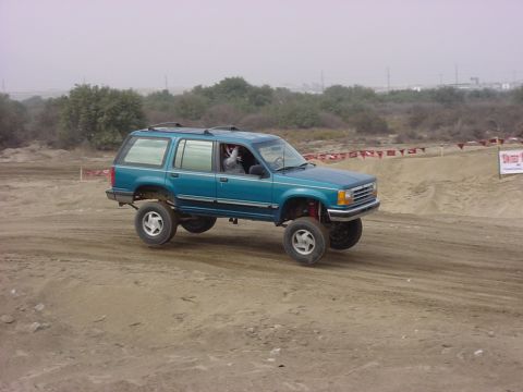 1984 Ford Bronco - A teal SUV with a lifted suspension navigates a dirt track, with a SUPERMOTORS watermark visible at the bottom right.