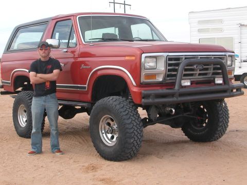 1984 Ford Bronco - A man stands next to a lifted red Ford Bronco with large off-road tires.