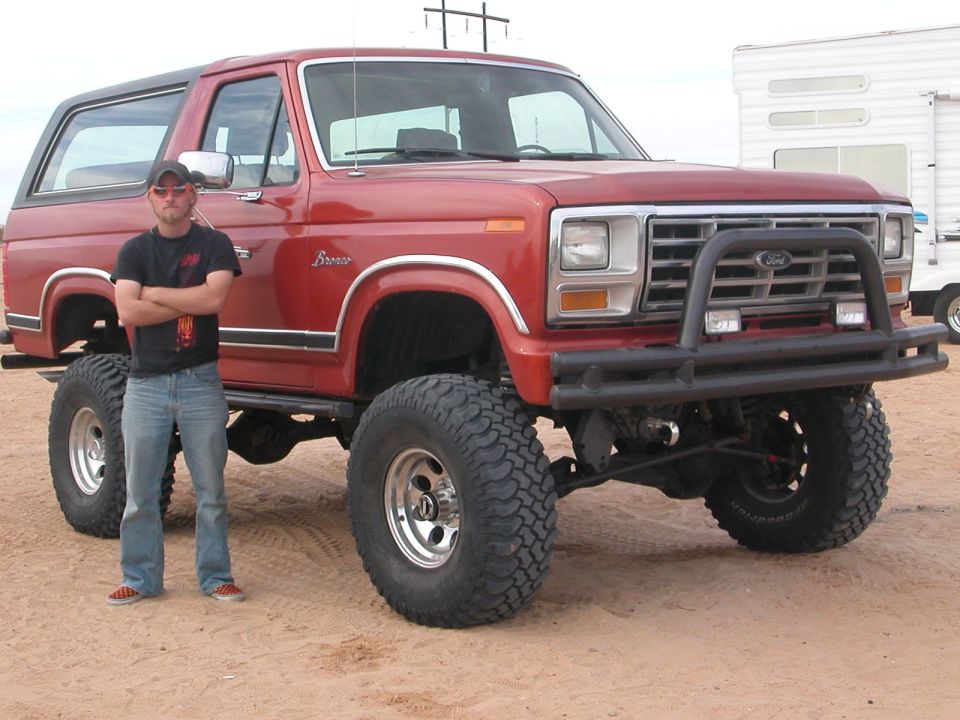 Red lifted Ford Bronco with a man standing beside it. Bronco badging visible