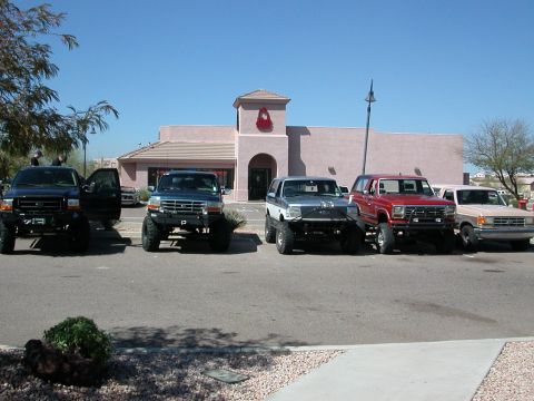 1984 Ford Bronco - Several Ford Broncos and F-Series trucks are lined up in a parking lot under a clear blue sky.