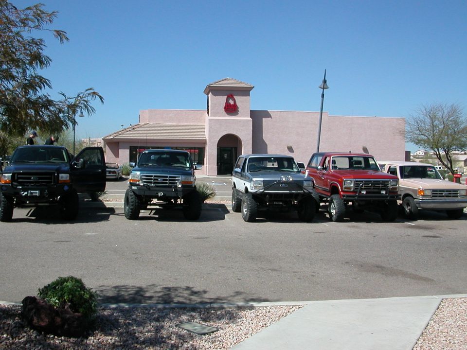 Five lifted Ford trucks and Broncos parked in front of a building. Blue, silver, red, and tan vehicles are visible