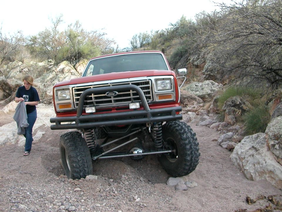 Red Ford pickup truck with a brush guard on a rocky trail. A woman stands to the left