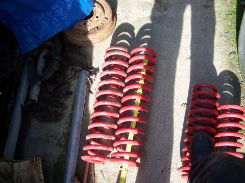 1996 Ford Bronco - Four red-painted coil springs laid out on a concrete driveway with a yellow tape measure stretched along one pair to compare free heights,…