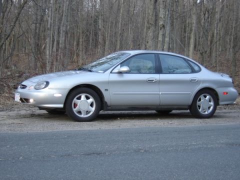 1999 Ford Taurus - Side view of a silver 1999 Ford Taurus parked on a roadside with bare trees behind it.