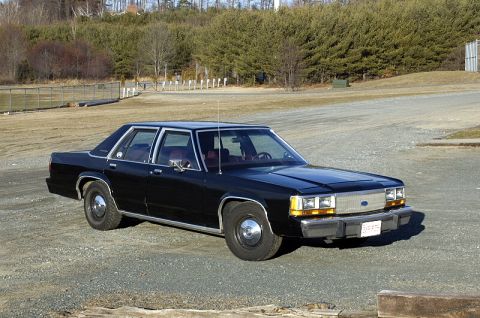 1991 Ford Crown Victoria - This black sedan is parked outdoors on a gravel surface with trees and a fence in the background.