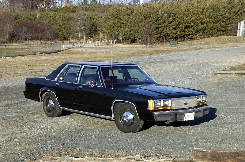 Black Ford Crown Victoria sedan with dog-dish hubcaps and whip antenna on gravel lot