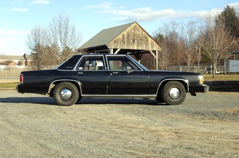 1991 Ford Crown Victoria - Side view of a black sedan parked outdoors on a gravel surface.