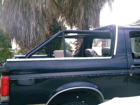 1989 Ford Bronco - The rear section of a dark blue SUV features a custom black roll cage, with palm trees in the background.
