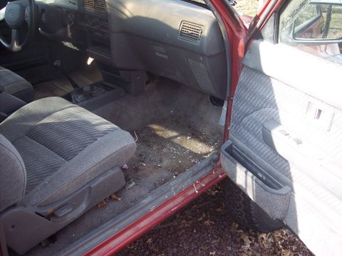 1995 Toyota Tacoma - The interior of a red truck shows a dirty floor and worn grey seats, with the driver's side door ajar.