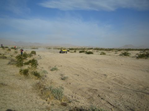 Yellow off-road buggy seen across a wide sandy desert with spectators