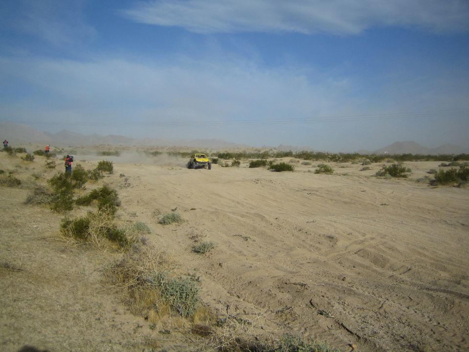 Yellow off-road buggy seen across a wide sandy desert with spectators