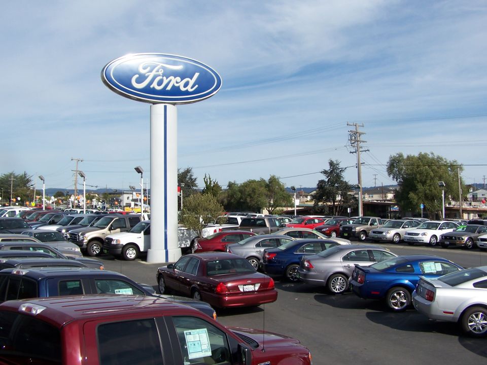 Ford dealership lot with a large Ford sign and many cars parked under a blue sky