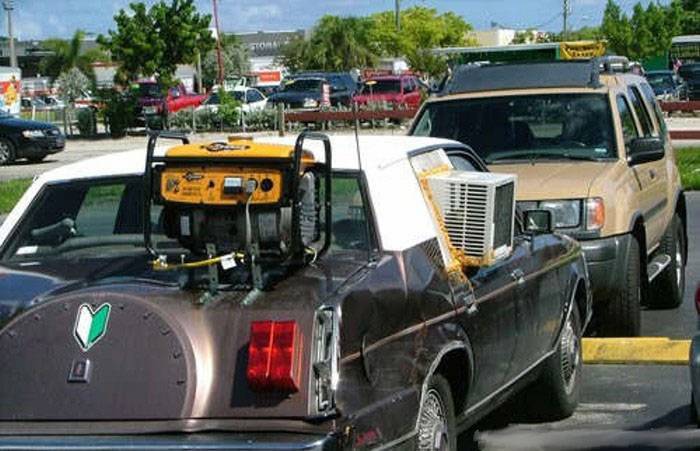 Brown 1970s sedan with a generator and window AC unit loaded on the trunk in a lot