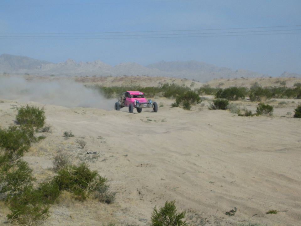 Pink off-road race buggy driving across open sandy desert with mountains behind