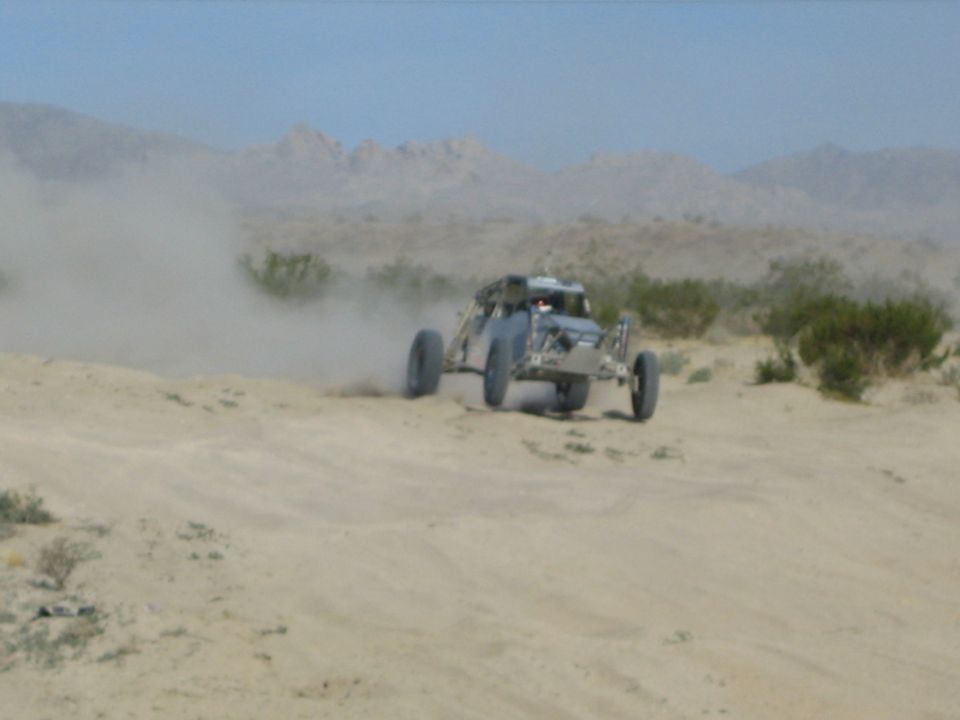 Silver off-road race buggy cresting a sandy desert rise trailing dust