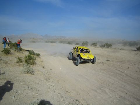 Yellow off-road race buggy kicking up dust on a desert trail with spectators watching
