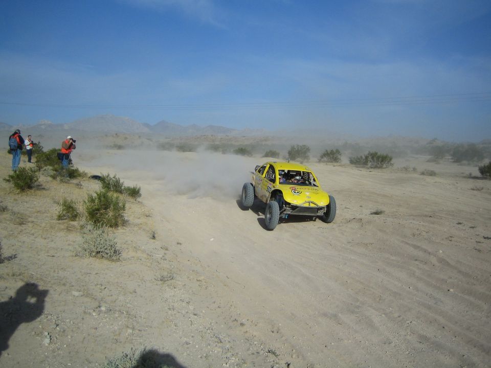 Yellow off-road race buggy kicking up dust on a desert trail with spectators watching