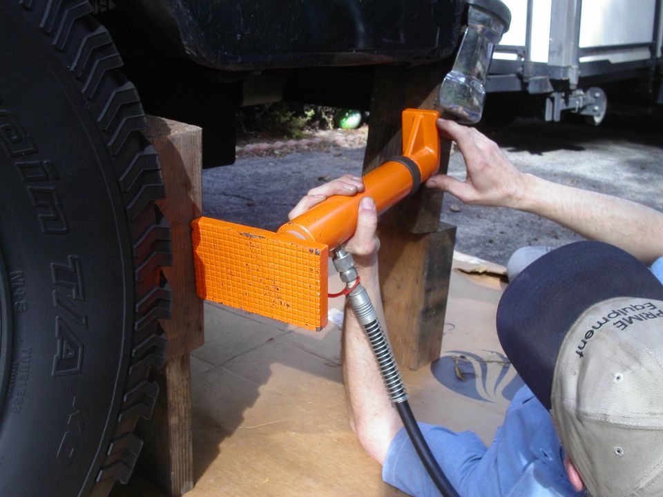 Person positioning an orange hydraulic ram on wood blocks under a vehicle beside a large tire