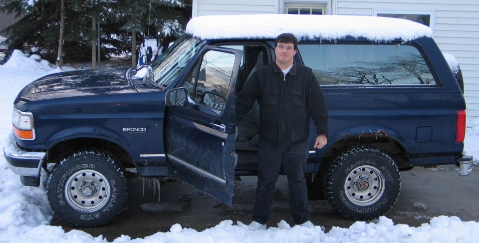 Man standing by open driver's door of dark blue Ford Bronco with snow-covered roof in winter driveway